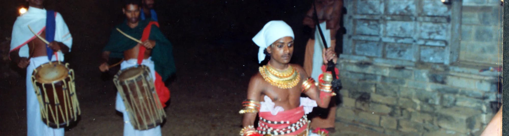 A costumed teyyam performer in headdress and ornaments standing at night beside two drummers playing chenda.