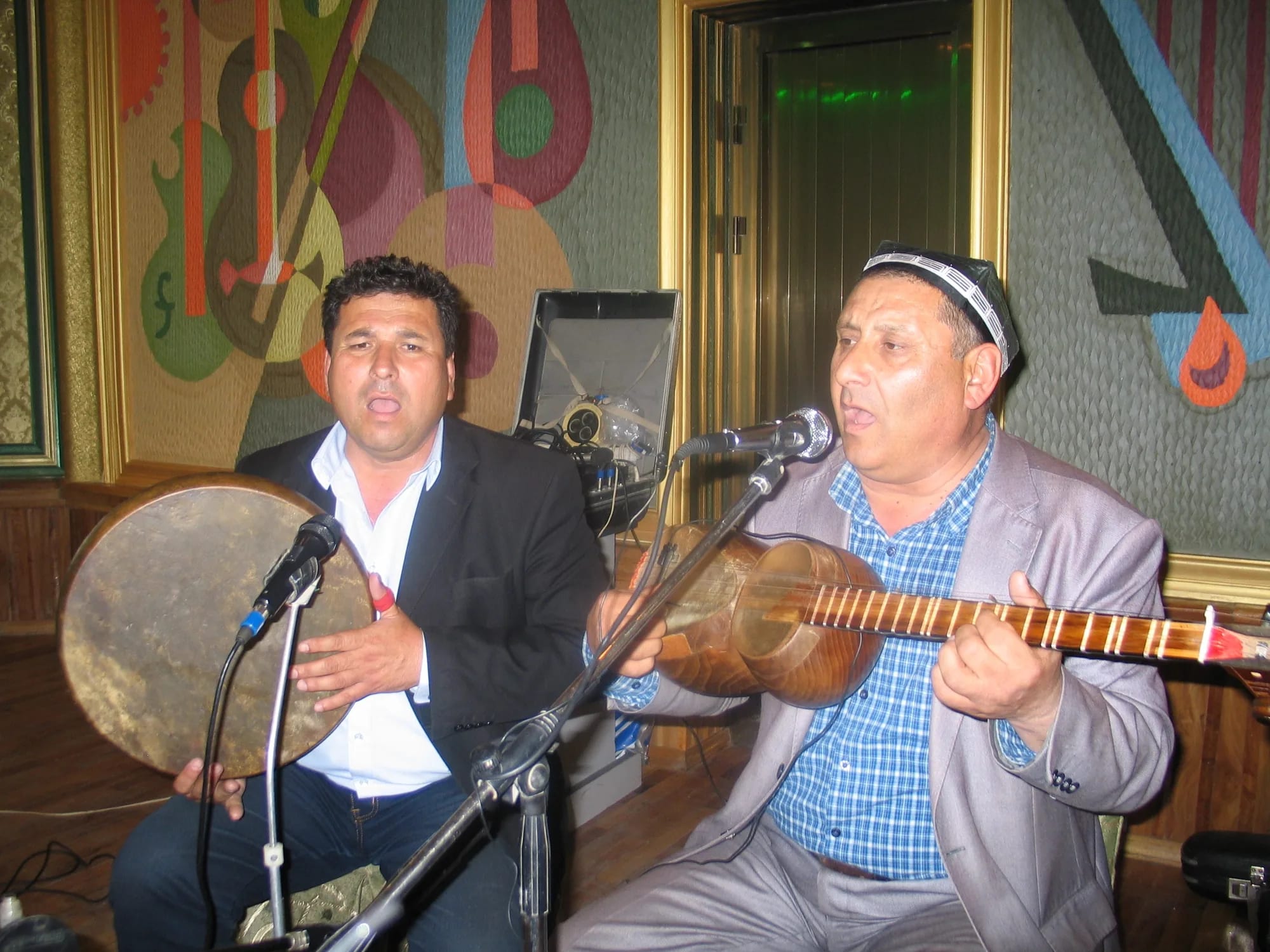 Two Tajik musicians performing at a microphone, one with a frame drum and one with a long-necked lute.