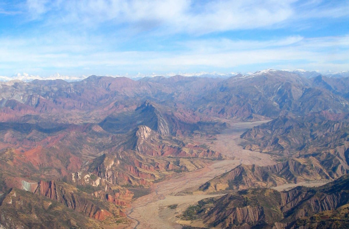An aerial view of eroded red and grey folded mountains cut by a wide braided riverbed, with snow peaks on the horizon.