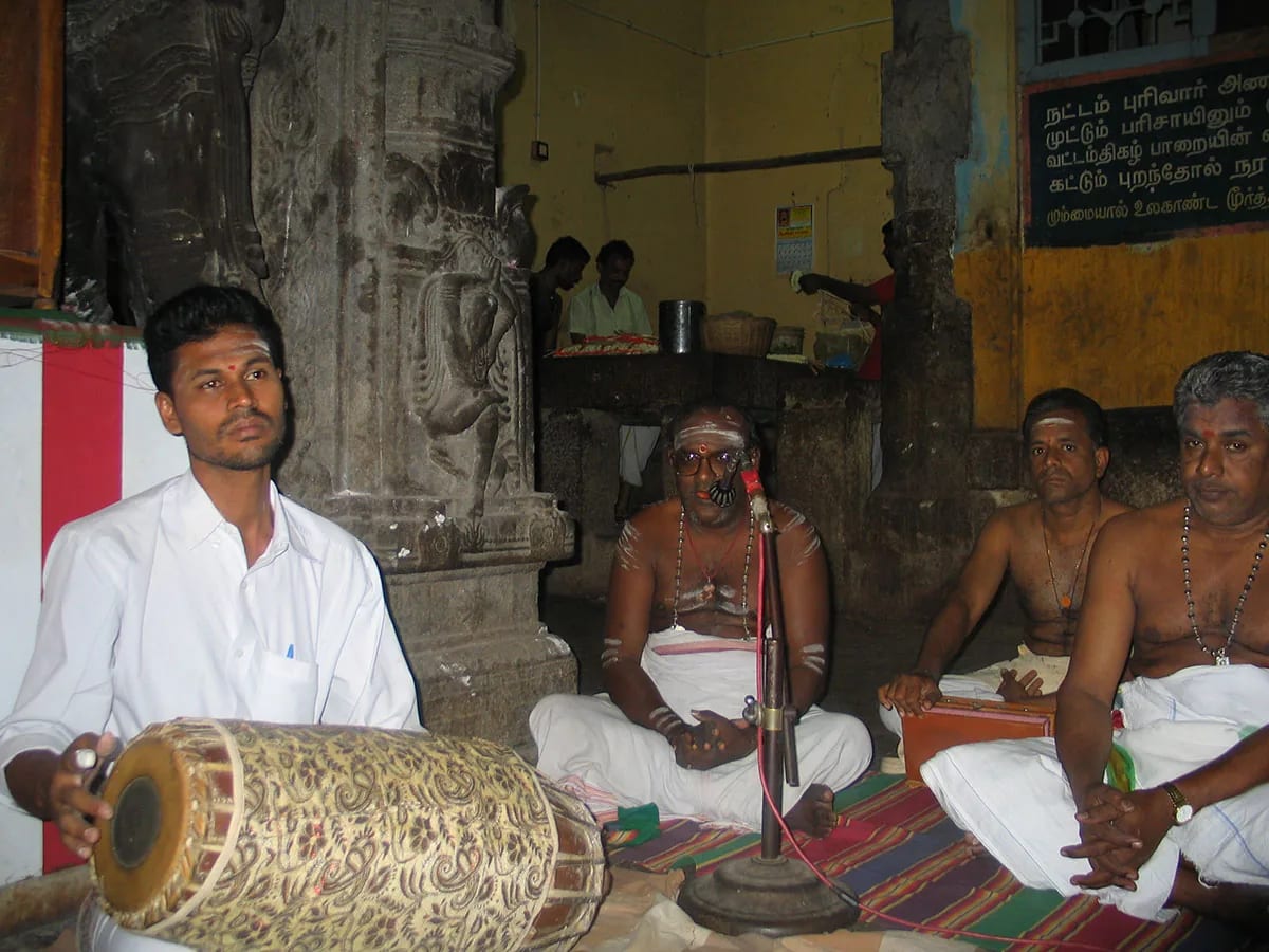 A young mridangam player and three singers seated on the floor of a temple hall before a carved stone pillar.