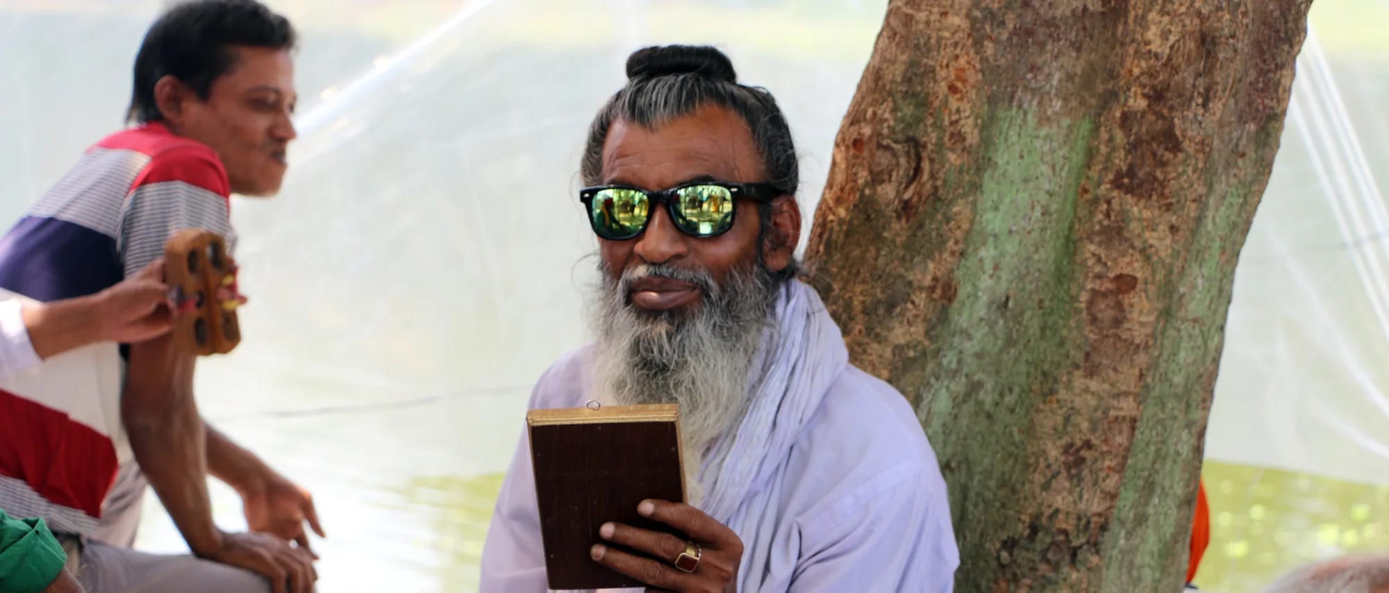 A bearded man in white, wearing mirrored sunglasses, reading from a small book beneath a tree.