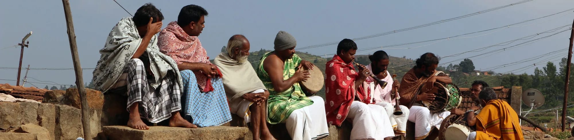 A row of Kota men and women seated together on a low stone wall, one holding a frame drum, hills behind them.